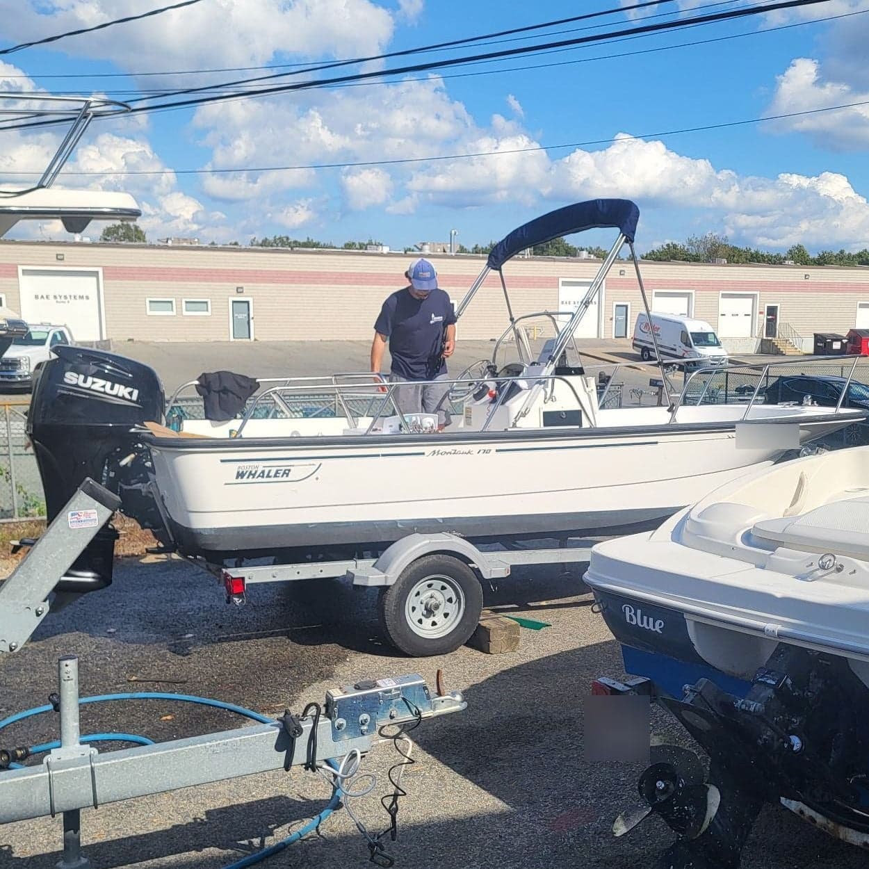 Man in a cap and navy shirt stands in a white Boston Whaler boat with a Suzuki outboard, on a trailer in a paved lot